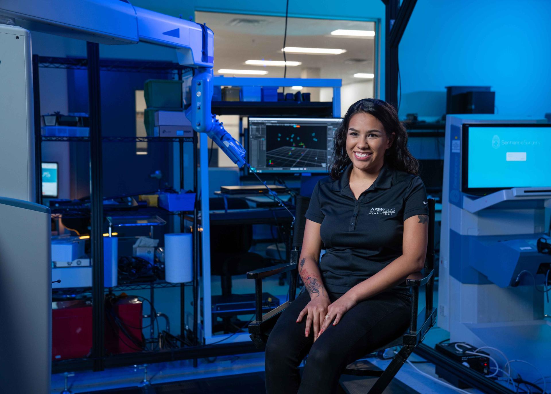 Smiling woman wearing an Asensus Surgical shirt in front of surgical robots